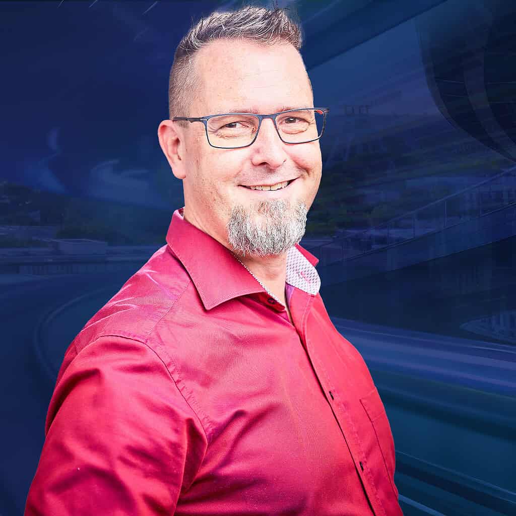 Ein Mann in einem auffälligen roten Hemd und einer Brille, lächelt in die Kamera vor einem blauen futuristischen Hintergrund.