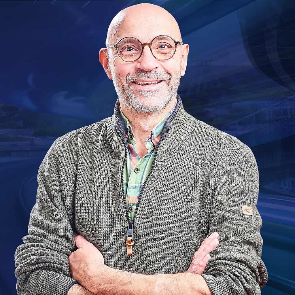 Ein Mann mit Glatze guckt mit verschränkten Armen und einem Lächeln in die Kamera.