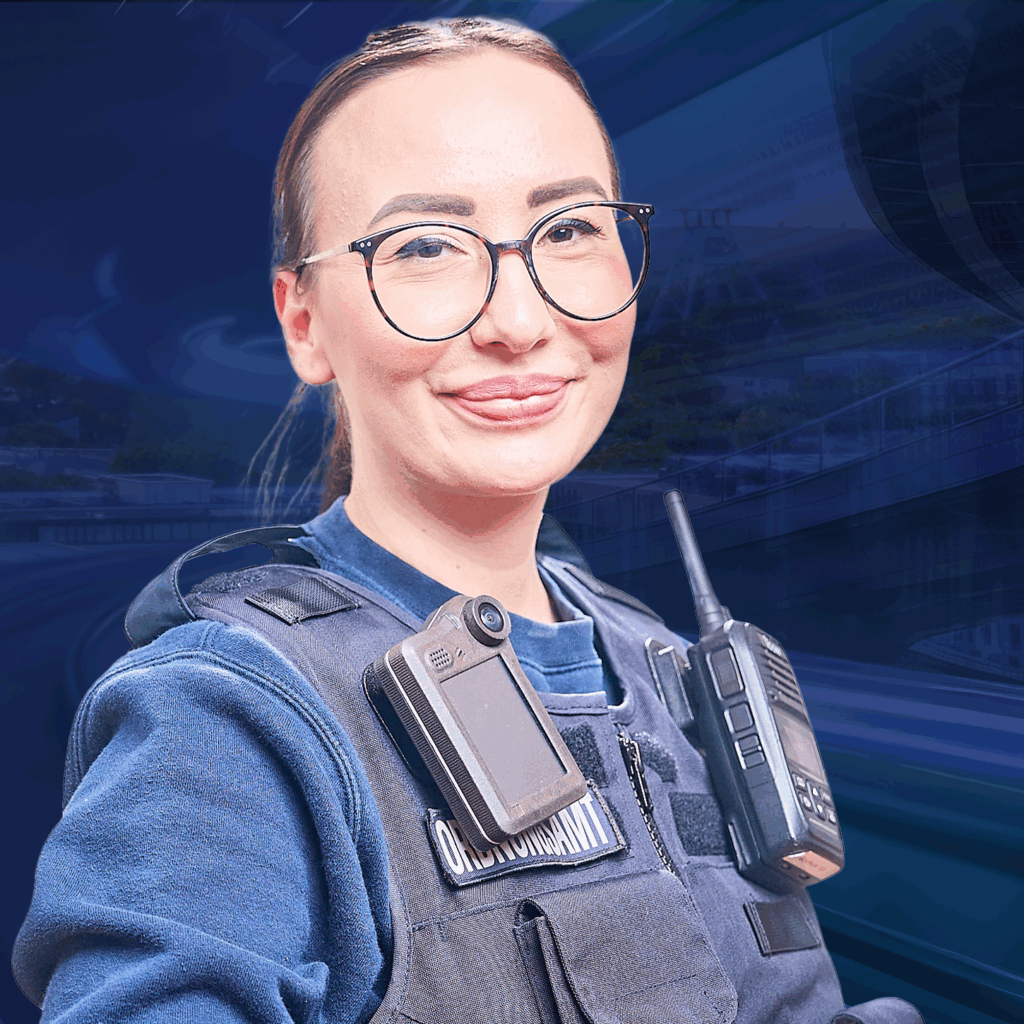 Eine lächelnde Frau in Uniform mit Funkgerät blickt selbstbewusst in die Kamera vor einem unscharfen Hintergrund.