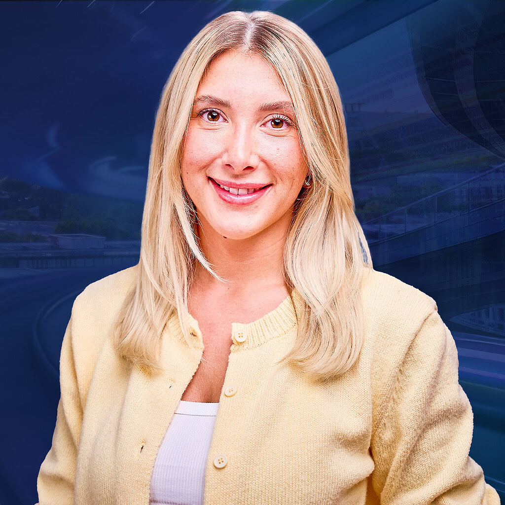 Eine lächelnde blonde Frau in gelber Strickjacke steht vor einem verschwommenen blauen Hintergrund.