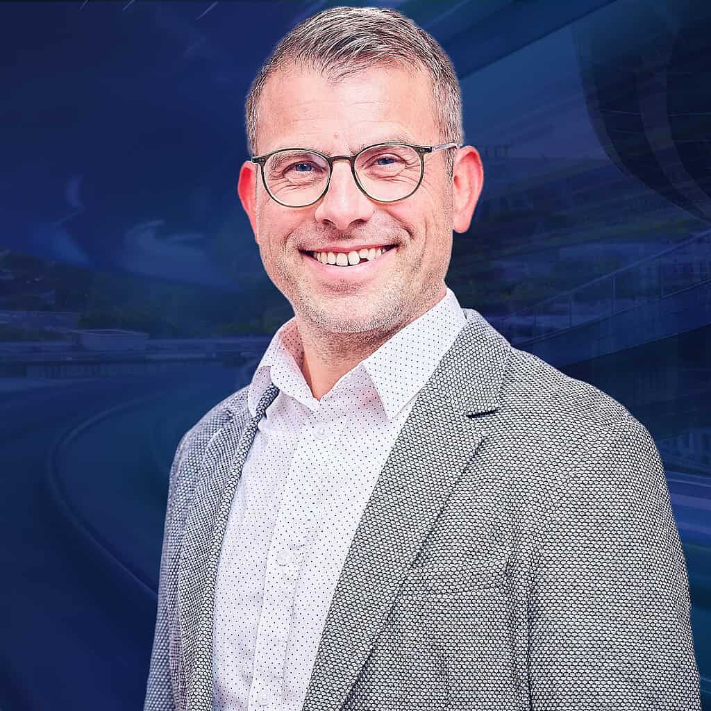 Ein lächelnder Mann in Brille und Anzug steht vor einem blauen Hintergrund mit futuristischen Details.