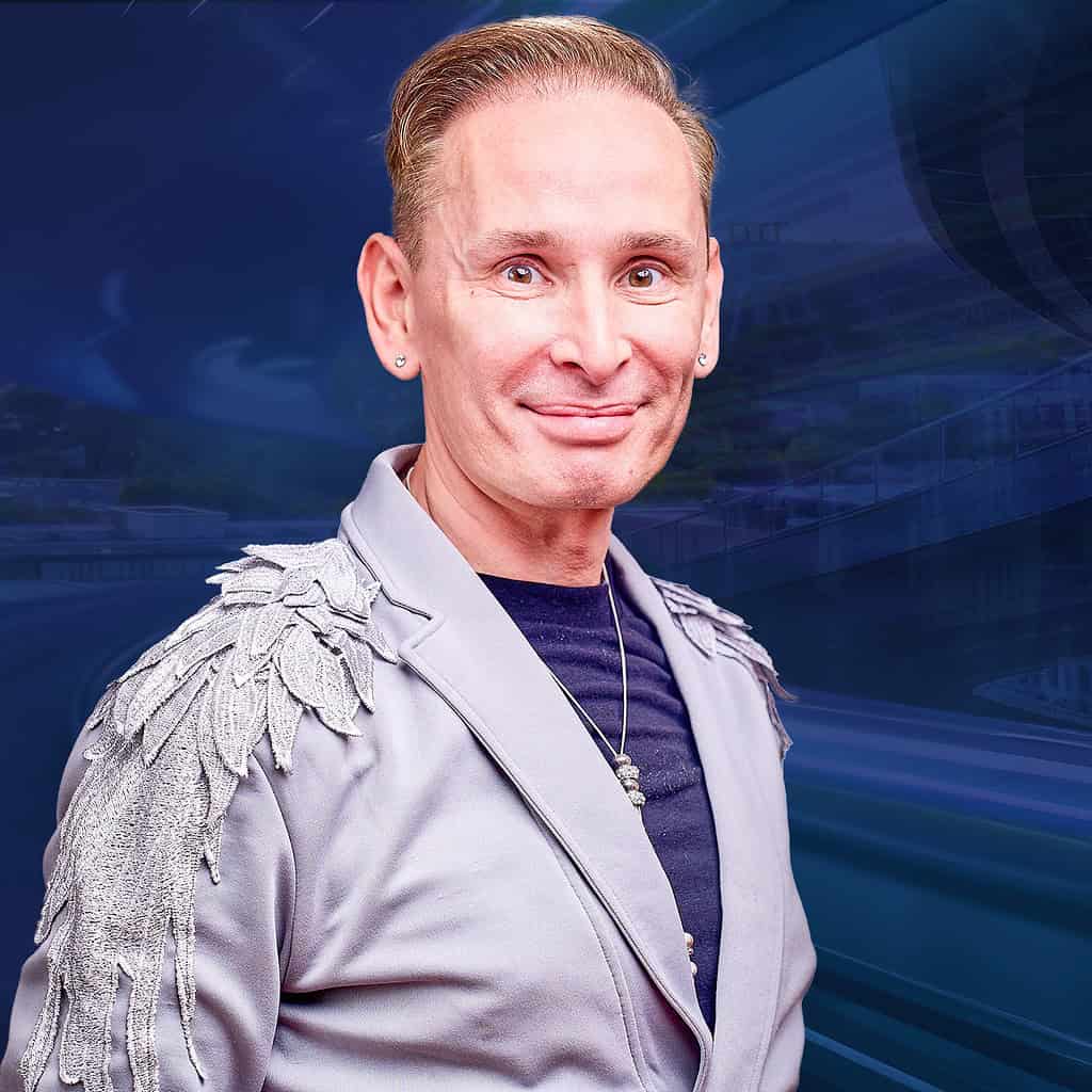 Ein lächelnder Mann trägt ein silbergraues Jackett mit dekorativen Schulterdetails vor einem blauen Hintergrund.